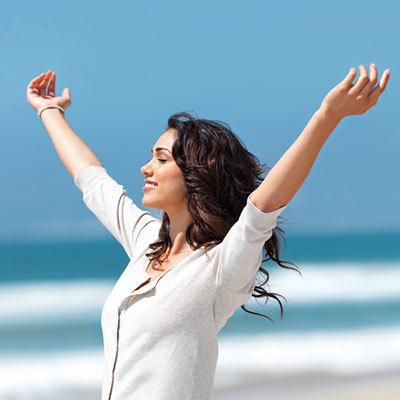 Image of a young woman on a beach with the ocean waves in the background. Her arms are raised wide and her smiling face is directed at ths sky